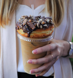Person holding iced coffee and a decadent chocolate doughnut at Deviant Doughnuts 